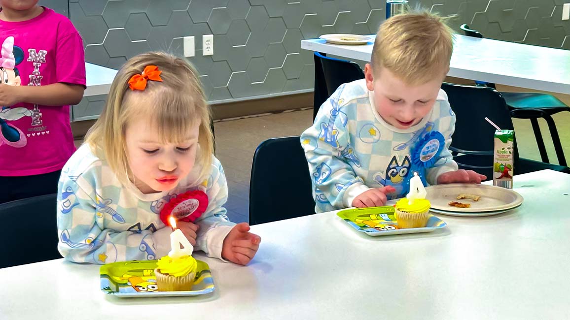 Two young children blow out candles on birthday cupcakes during a celebration at Science Museum Oklahoma, surrounded by friends in a decorated party room.