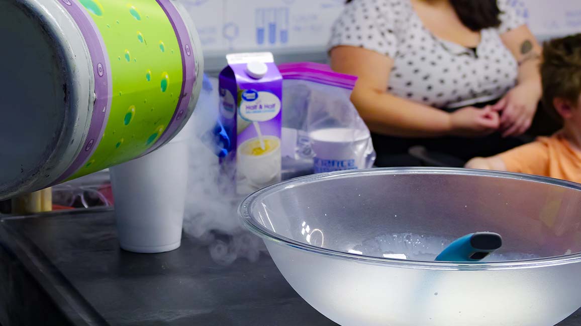 A Science Museum Oklahoma staff member pours liquid nitrogen into a mixing bowl during a live birthday party demonstration, with families watching in amazement.