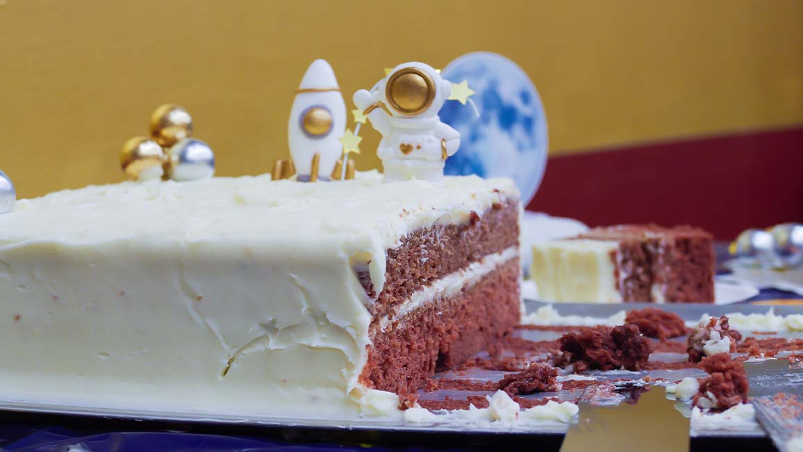 A partially eaten space-themed red velvet birthday cake decorated with planets, a rocket, and an astronaut sits on a party table at Science Museum Oklahoma.