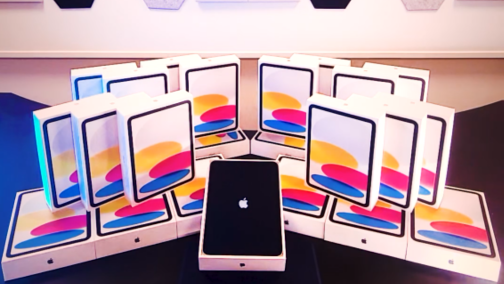 A group of boxed Apple iPads arranged in a semi-circle on a table, with one unboxed iPad standing upright in the center displaying the Apple logo.