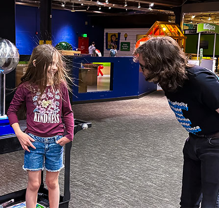 A science Museum Oklahoma Employee bends down to talk to a gir who is experiencing electrified hair in an experiment. 