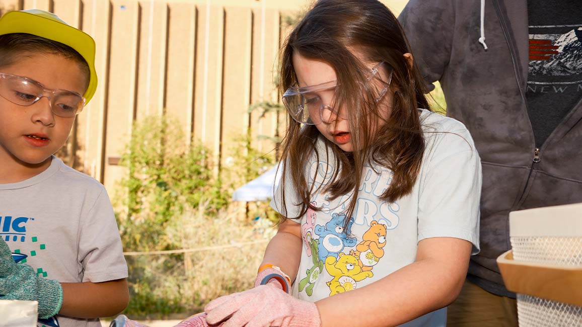 A young girl wearing safety gear cuts a soda can during an outdoor hands-on science activity at Science Museum Oklahoma while another child observes at DiscoverFest.