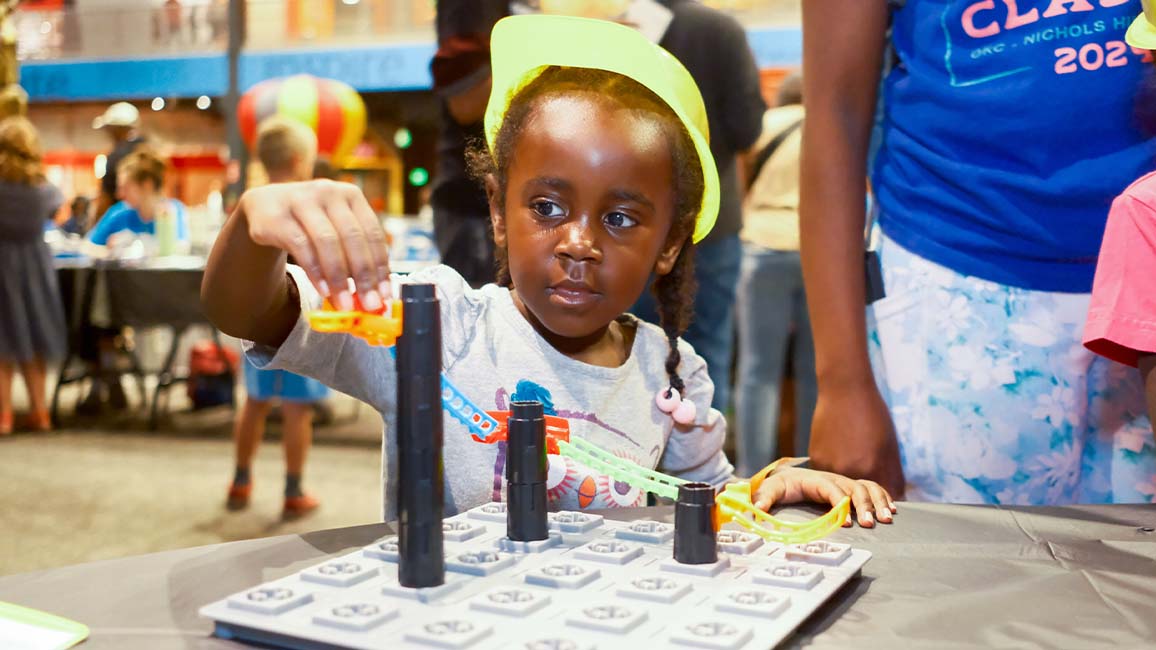 A young girl wearing a yellow hard hat builds a roller coaster track during a hands-on STEM activity at Science Museum Oklahoma, exploring engineering through play at DiscoverFest.