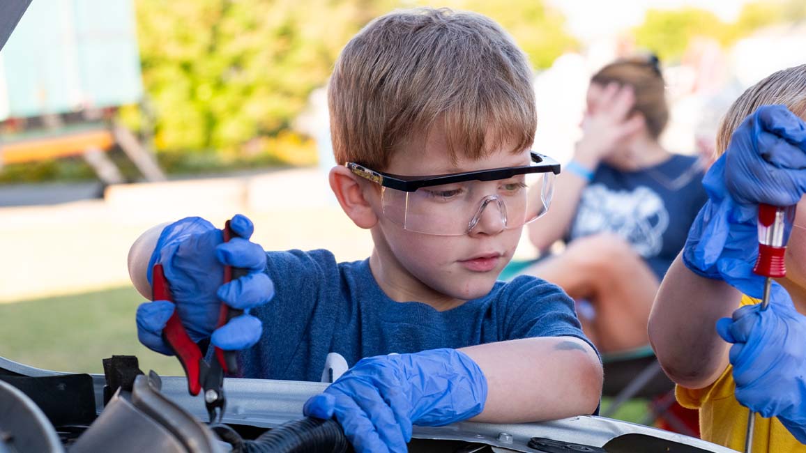 A young boy wearing safety goggles and gloves uses tools to explore car mechanics during a hands-on STEM activity at Science Museum Oklahom at DiscoverFEST.