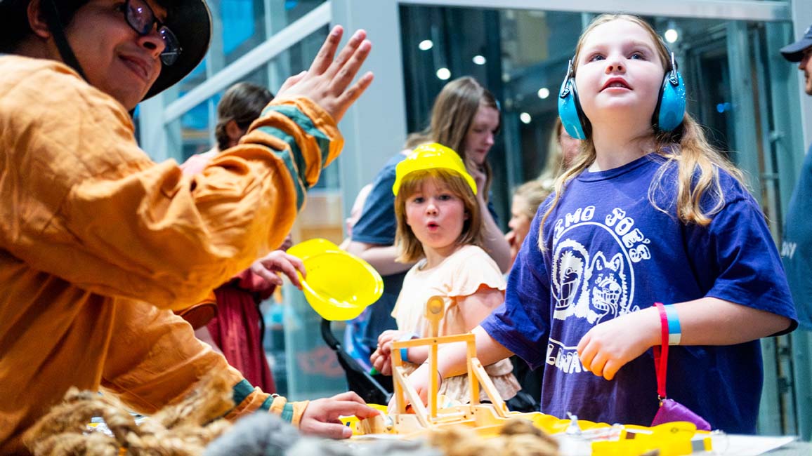 Children engage in an interactive hands-on STEM activity with a costumed educator at Science Museum Oklahoma, exploring engineering with ear protection and curiosity AT DiscoverFest.