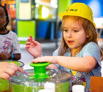 A young child wearing a hard hat engages in a hands-on science activity at Science Museum Oklahoma, exploring art and motion through a colorful spin experiment at DiscoverFest.
