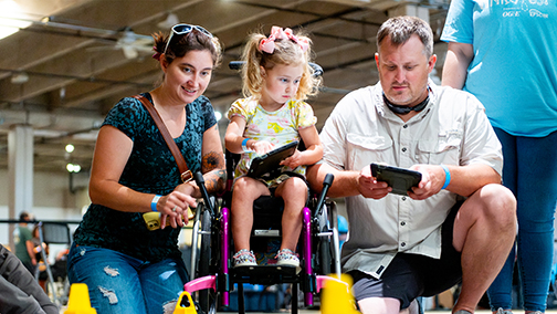 A family explores a hands-on STEM activity at Science Museum Oklahoma, with a young child in a wheelchair using a tablet to navigate a robotics course.