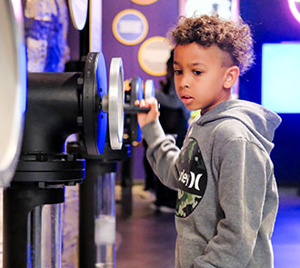 Young boy in a gray hoodie exploring a hands-on exhibit at Science Museum Oklahoma, turning a mechanical wheel in a brightly lit gallery.
