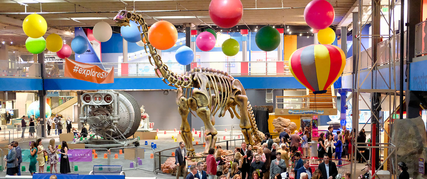 Guests enjoy an event called Elemental Ball, a fundraiser at Science Museum Oklahoma, surrounded by colorful balloons, a towering dinosaur skeleton, and interactive exhibits in the main museum which is available for rental.