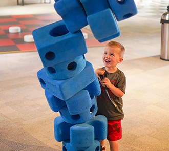 A young boy smiles at a towering stack of oversized foam blocks in the Big Game Theory exhibit at Science Museum Oklahoma.