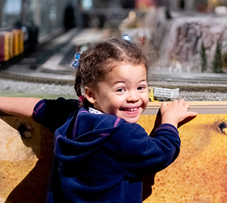A young child smiles while exploring a detailed model train exhibit at Science Museum Oklahoma’s Tiny Tracks.
