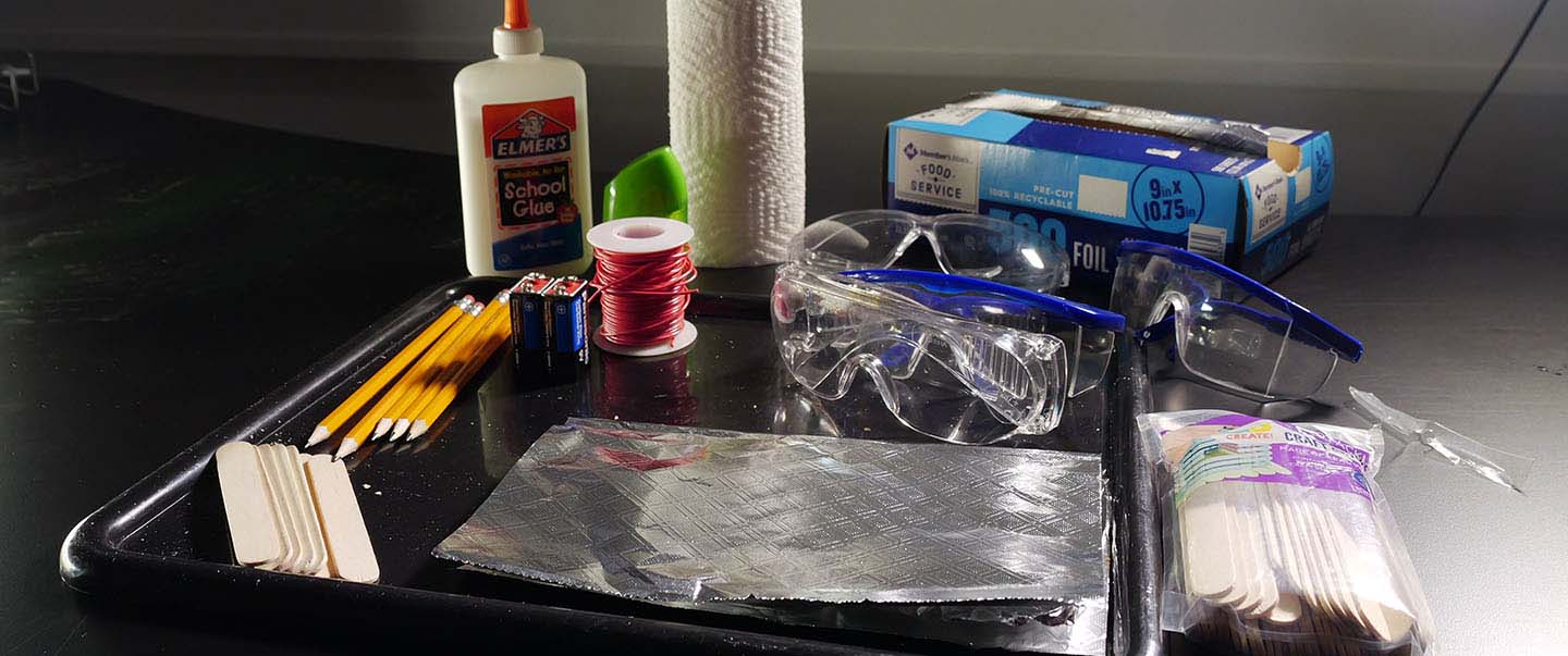 A collection of hands-on science materials including goggles, foil, glue, wire, and batteries on a black table for a family STEM activity at Science Museum Oklahoma.