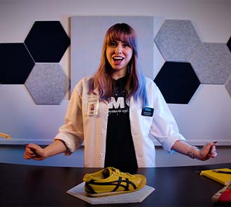 A Science Museum Oklahoma educator in a white lab coat presents a STEM activity on a table with a yellow sneaker and science equipment for families at home.