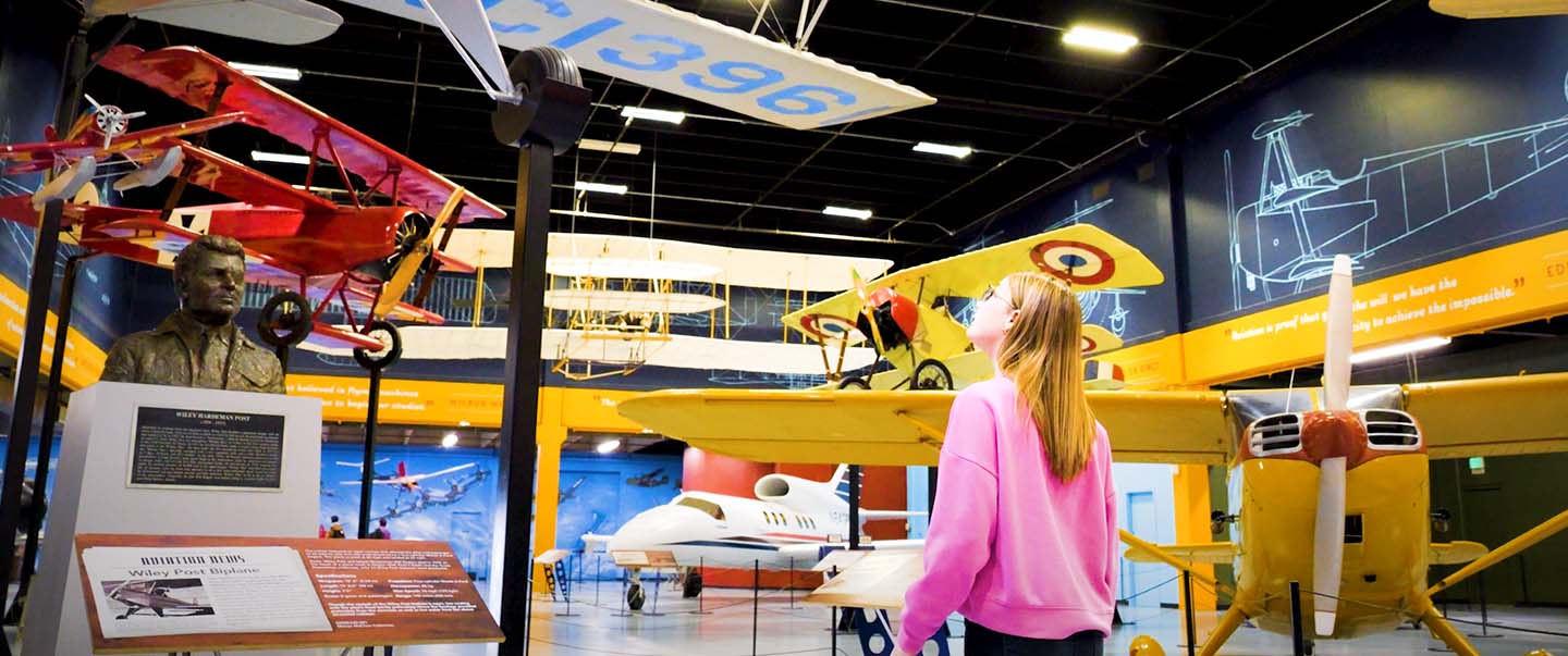 A young girl looks up at colorful vintage aircraft hanging from the ceiling in Science Museum Oklahoma's aviation exhibit.