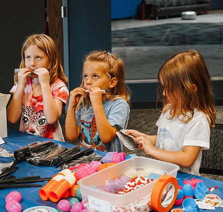 Three young girls sit at a craft table in Science Museum Oklahoma, experimenting with small plastic instruments while colorful supplies like beads, stickers, and plastic eggs are spread out in front of them.