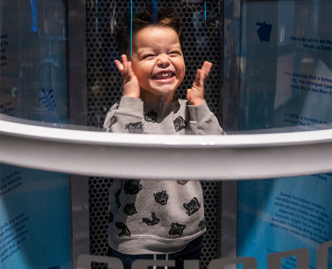 A young child grins inside the Oklahoma Museum Network’s wind tunnel at Science Museum Oklahoma, experiencing hands-on weather science.