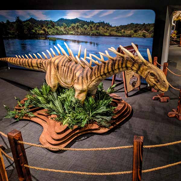 Life-sized dinosaur model with dramatic spikes displayed at Science Museum Oklahoma’s Expedition Dinosaur exhibit, surrounded by plants and roped barriers with a scenic prehistoric backdrop.