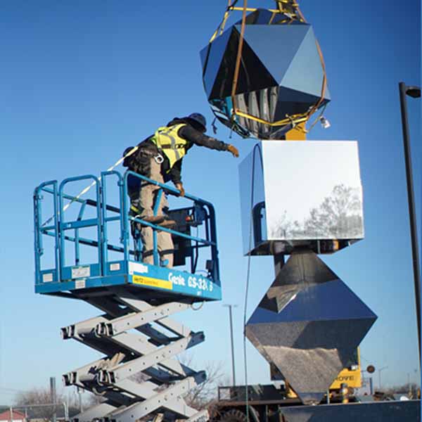 Worker in a safety harness using a scissor lift to install or repair a mirrored geometric sculpture outdoors at Science Museum Oklahoma, with a crane assisting above.
