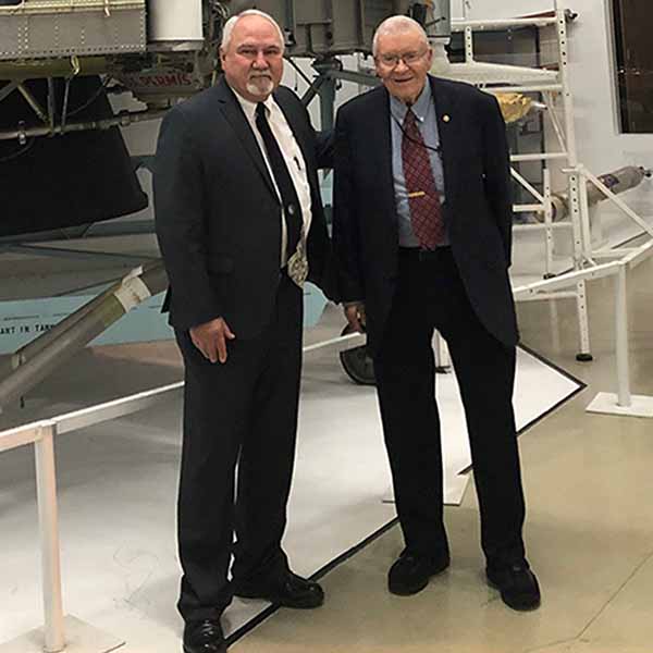 Two men in suits stand in front of a large spacecraft component inside Science Museum Oklahoma’s aviation and space exhibit.