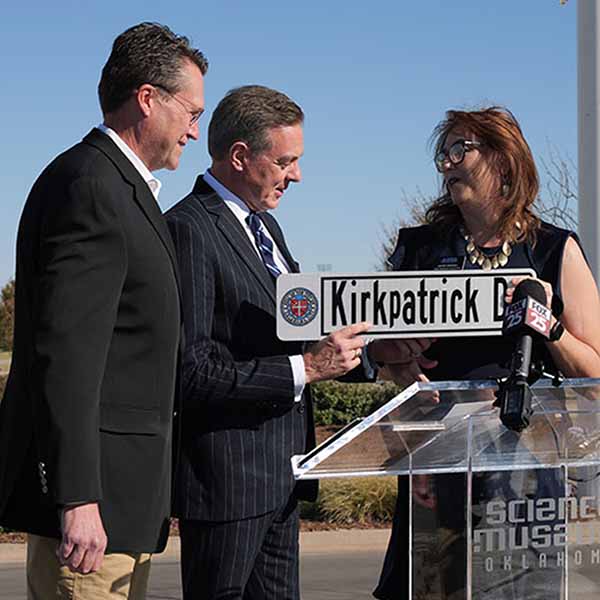 Three individuals standing at a podium outside Science Museum Oklahoma during a street renaming ceremony, holding a "Kirkpatrick Dr" sign with microphones nearby, including one labeled FOX 25.