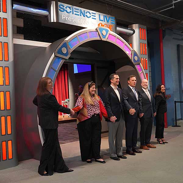 A group of 6 adults hold hands to activate a science demonstration at the entrance to the recently opened Science Live Auditorium.