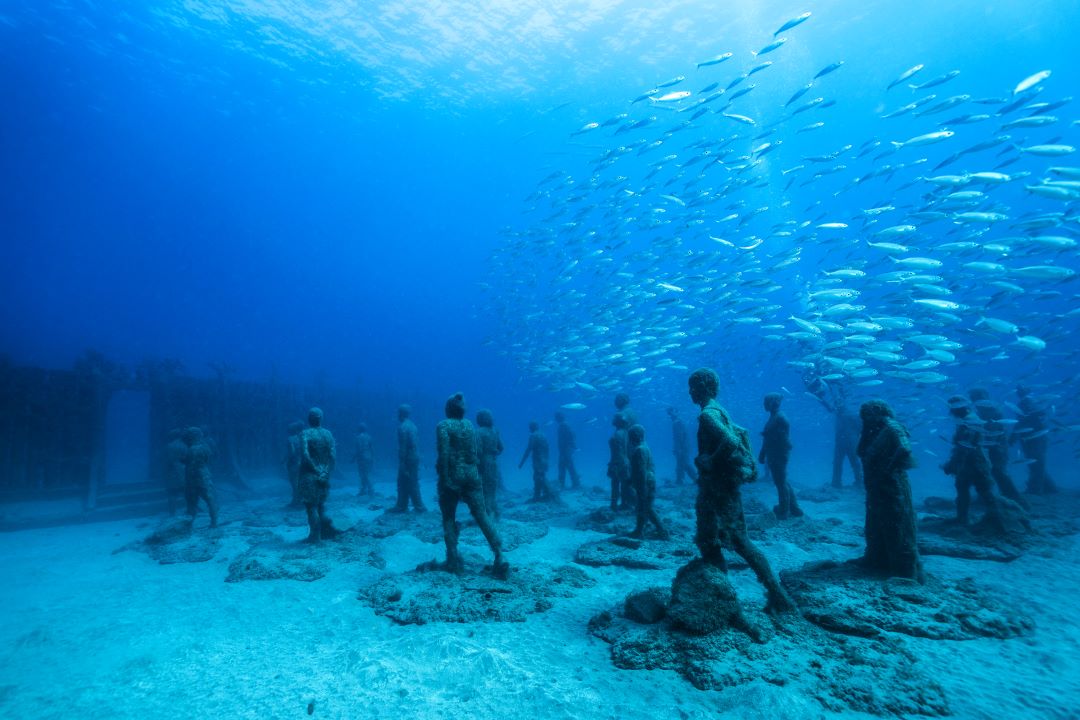Underwater sculpture installation titled "Crossing the Rubicon" by Jason deCaires Taylor, featuring life-sized human figures standing on the ocean floor near a school of fish, surrounded by deep blue water. Smart Space exhibit at Science Museum Oklahoma