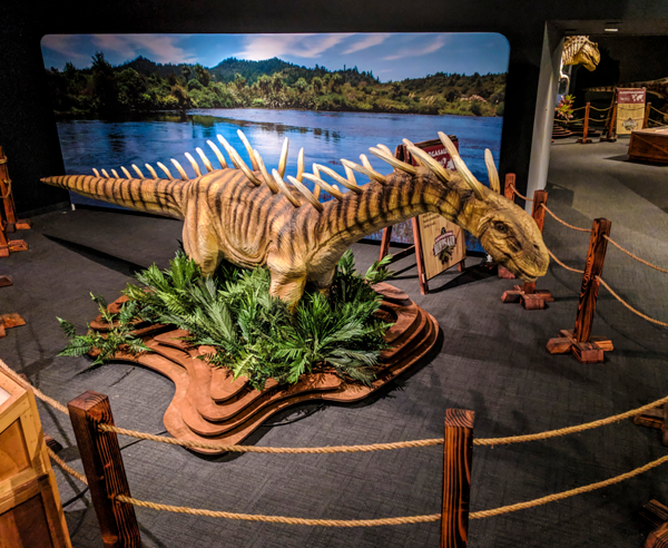 Life-sized dinosaur model with dramatic spikes displayed at Science Museum Oklahoma’s Expedition Dinosaur exhibit, surrounded by plants and roped barriers with a scenic prehistoric backdrop.