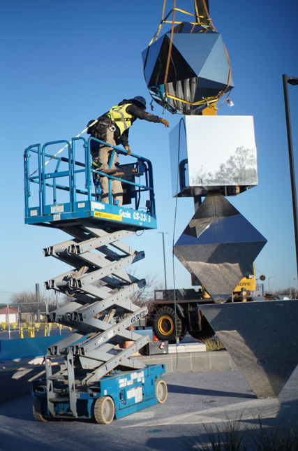 Worker in a safety harness using a scissor lift to install or repair a mirrored geometric sculpture outdoors at Science Museum Oklahoma, with a crane assisting above.