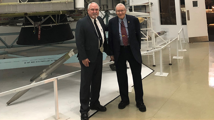 Two men in suits stand in front of a large spacecraft component inside Science Museum Oklahoma’s aviation and space exhibit.