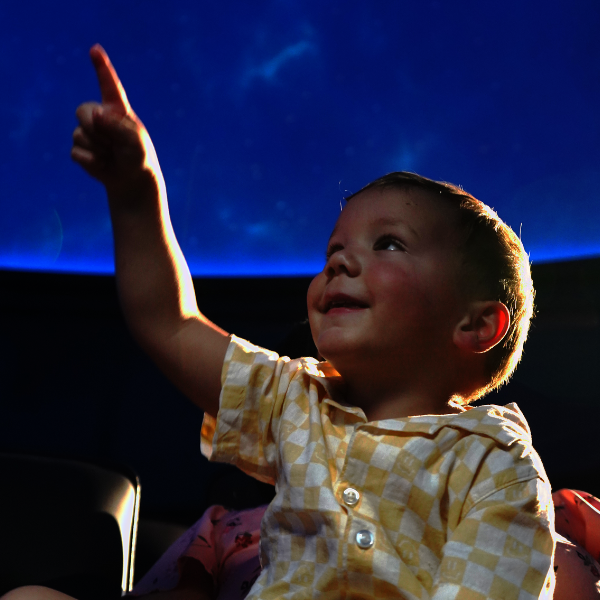 Young child smiling and pointing upward while seated inside a planetarium, illuminated by the glow of a starry sky projection overhead. Love's Planetarium Grand Opening