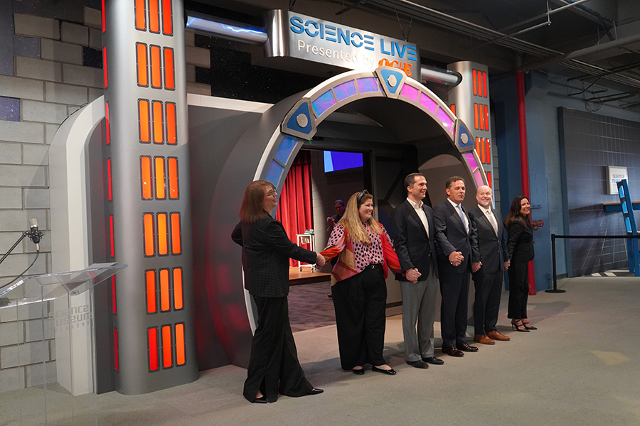Group of people holding hands and smiling in front of the newly unveiled Science Live theater entrance at Science Museum Oklahoma, presented by OG&E, during a ribbon-cutting or grand opening event.