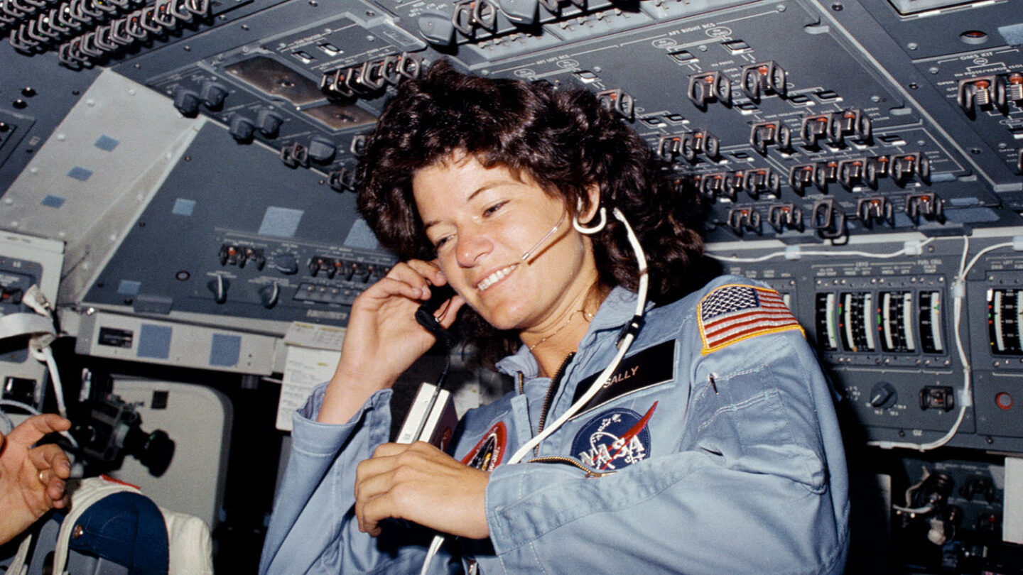 Astronaut Sally Ride inside the space shuttle cockpit wearing a NASA flight suit and headset, surrounded by control panels during her historic mission as the first American woman in space.
