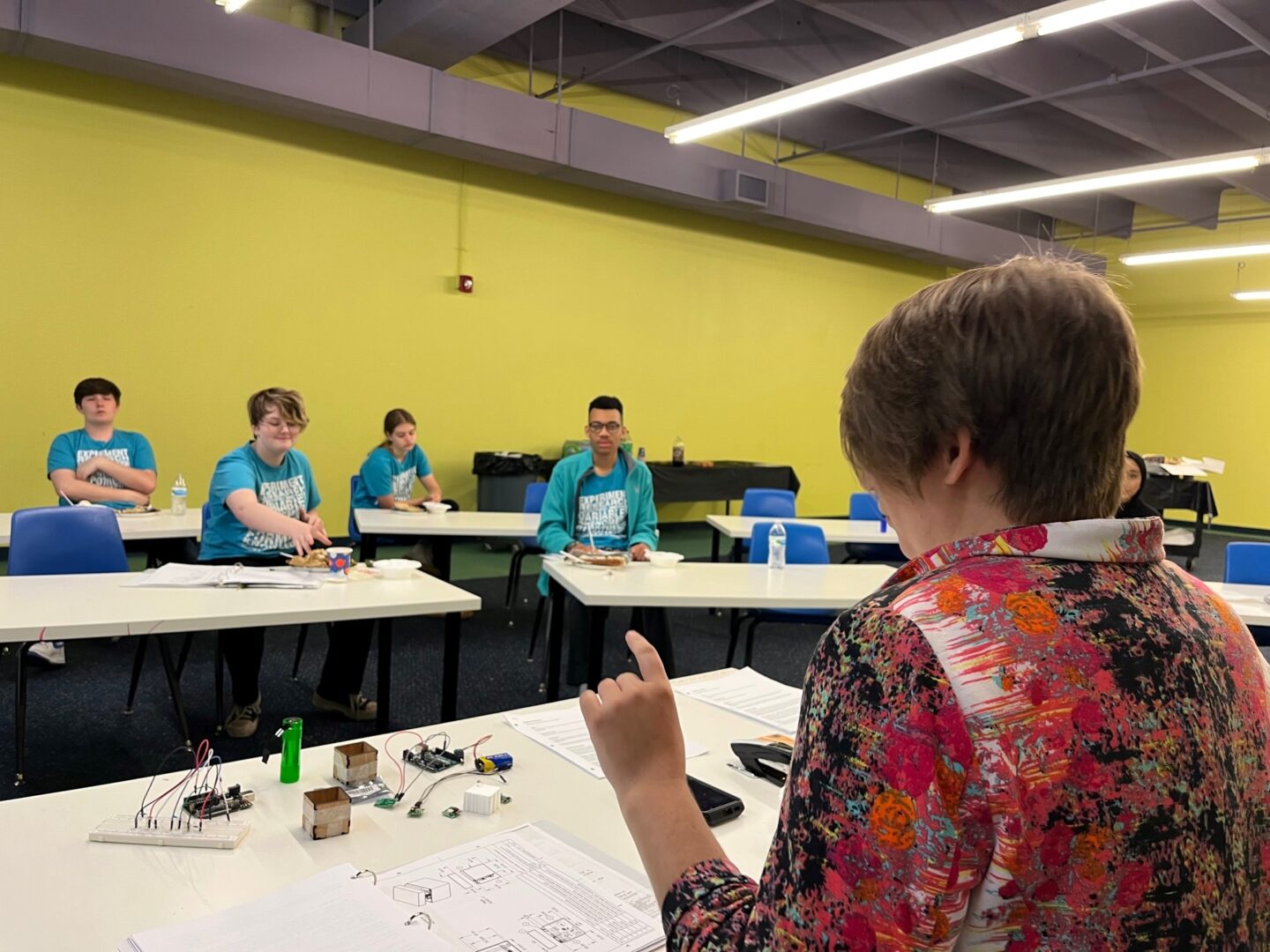 A teacher or instructor in a colorful floral shirt leads a classroom session with a group of teens seated at white desks. The students, wearing matching teal shirts with text that appears to say 'EXPERIMENT GALAXY' and 'NASA,' listen attentively. Various electronic components and circuit boards are laid out on the front table, along with technical diagrams, indicating a STEM-focused activity or workshop.