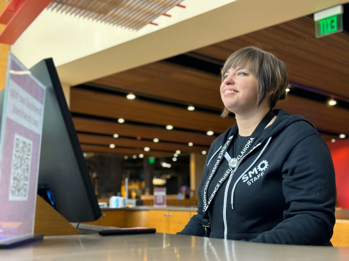 Science Museum Oklahoma staff member smiling at a front desk computer station, wearing a branded SMO hoodie and lanyard, with a warmly lit modern interior in the background.