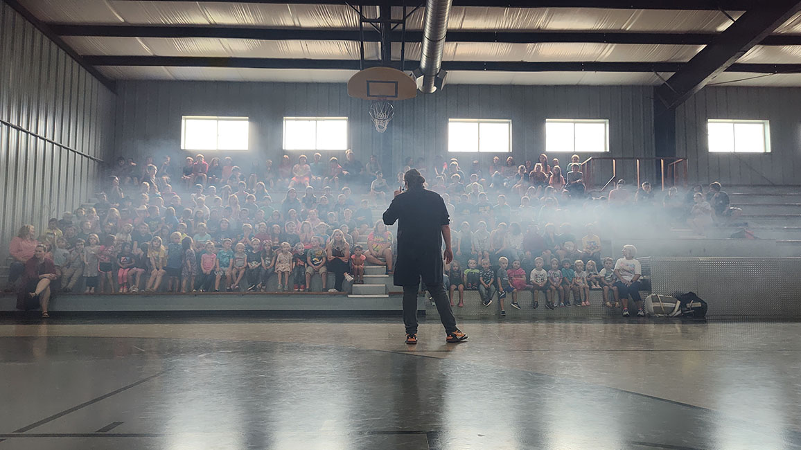 A man stands in front of a crowd of students at a rural school during a science presentation from Science Museum Oklahoma.
