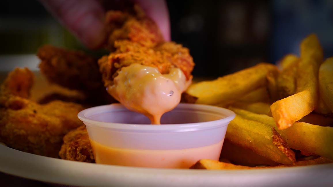 Hand-breaded chicken tenders and fries from Pavlov’s Café at Science Museum Oklahoma, with a tender being dipped in signature sauce.