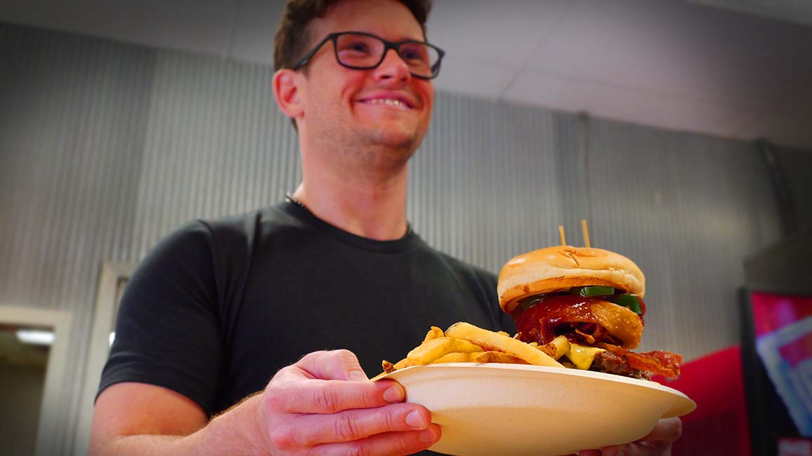 A smiling man holds a plate with a towering cheeseburger and golden fries at Pavlov’s Café inside Science Museum Oklahoma.