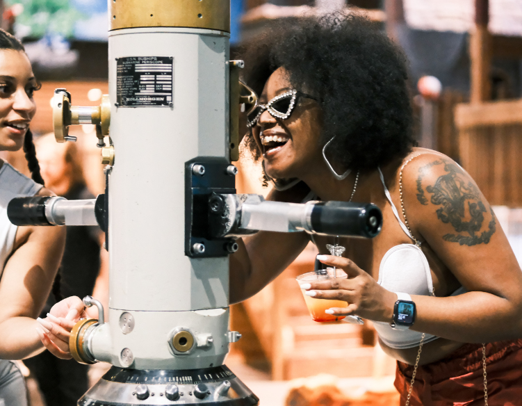 A woman wearing sparkly cat-eye sunglasses laughs while looking through a telescope at Science Museum Oklahoma’s SMO21+ event. She holds a cocktail in one hand and wears a white top and smartwatch.