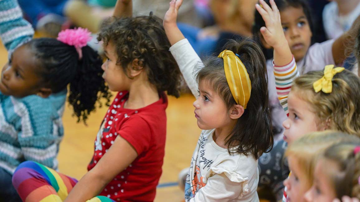 Young children at a Science Museum Oklahoma outreach excitedly raise their hands during an interactive science assembly in a school gym.