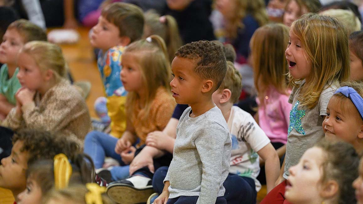 Children sit closely together on a gym floor watching a science demonstration from Science Museum Oklahoma’s STEAM Live program for schools.