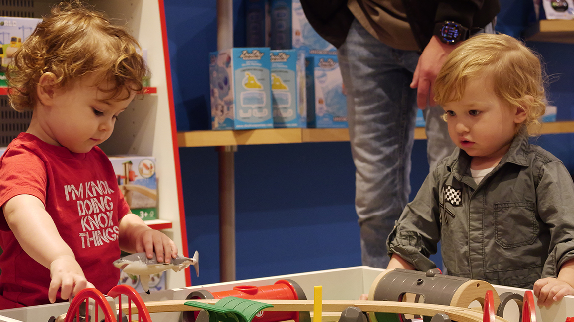 Two toddlers interact with a hands-on BRIO train table, one of the many interactive STEM play areas inside the Science Shop.