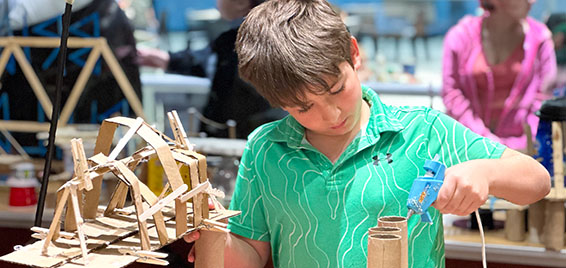 A child builds a cardboard structure using a hot glue gun in the Tinkering Garage at Science Museum Oklahoma, a hands-on makerspace for STEM creativity.
