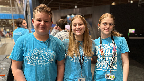 Three teen apprentices wearing blue Science Museum Oklahoma shirts smile for the camera inside the museum, with interactive exhibits and other visitors in the background.