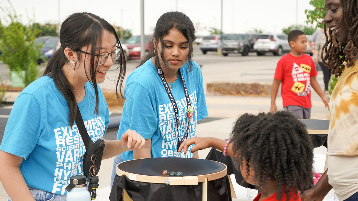 Two teen apprentices wearing blue Science Museum Oklahoma shirts demonstrate a marble activity to a child during an outdoor event, as families look on.