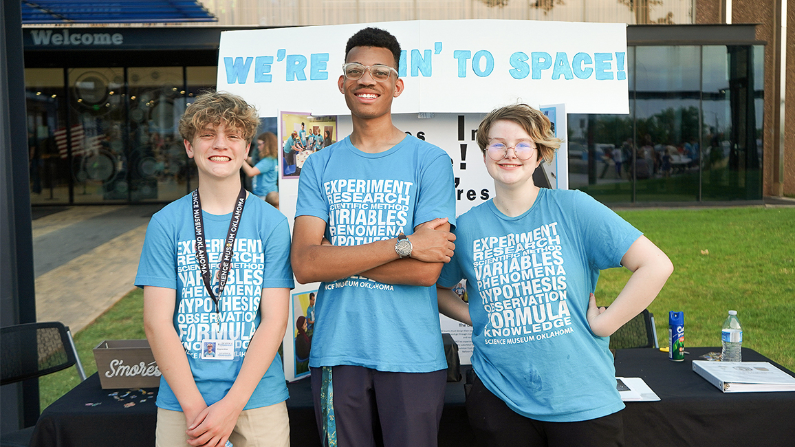 Three teen apprentices in blue Science Museum Oklahoma shirts stand smiling in front of a display table with a sign that reads “We’re Goin’ to Space!”