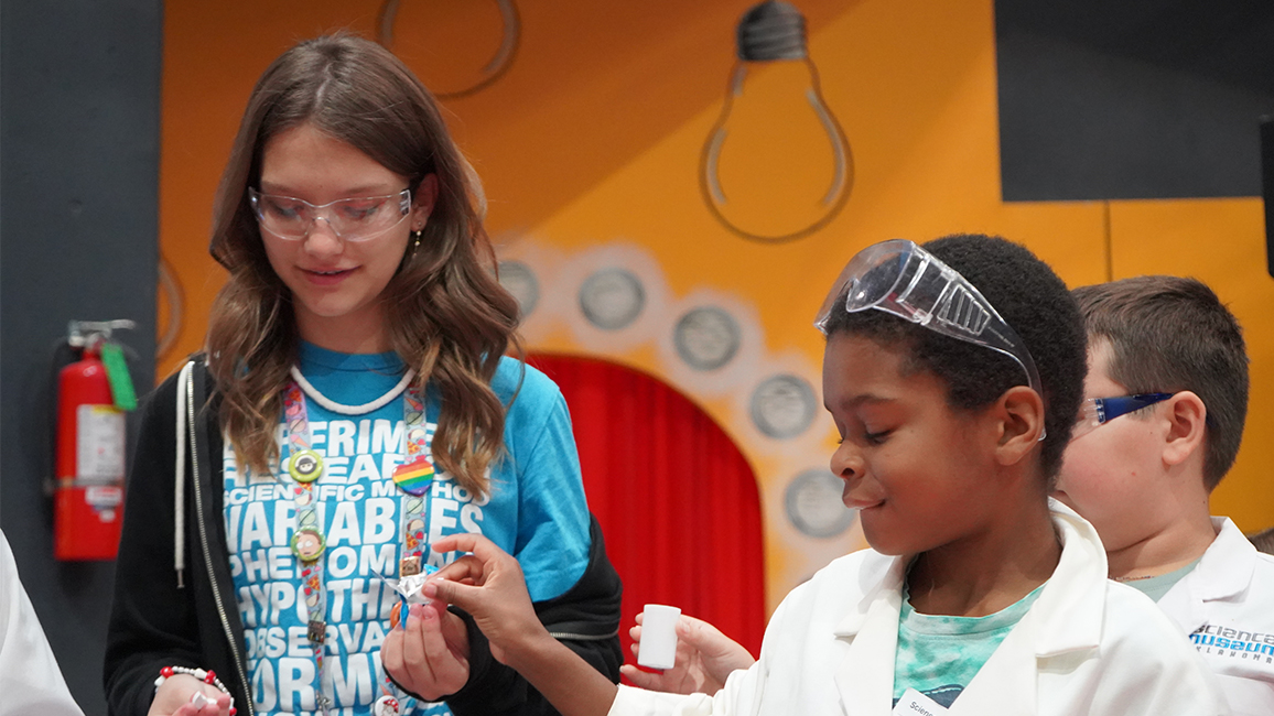 A teen apprentice wearing a blue Science Museum Oklahoma shirt and safety goggles helps two young children in lab coats with a science experiment inside a colorful museum exhibit area.
