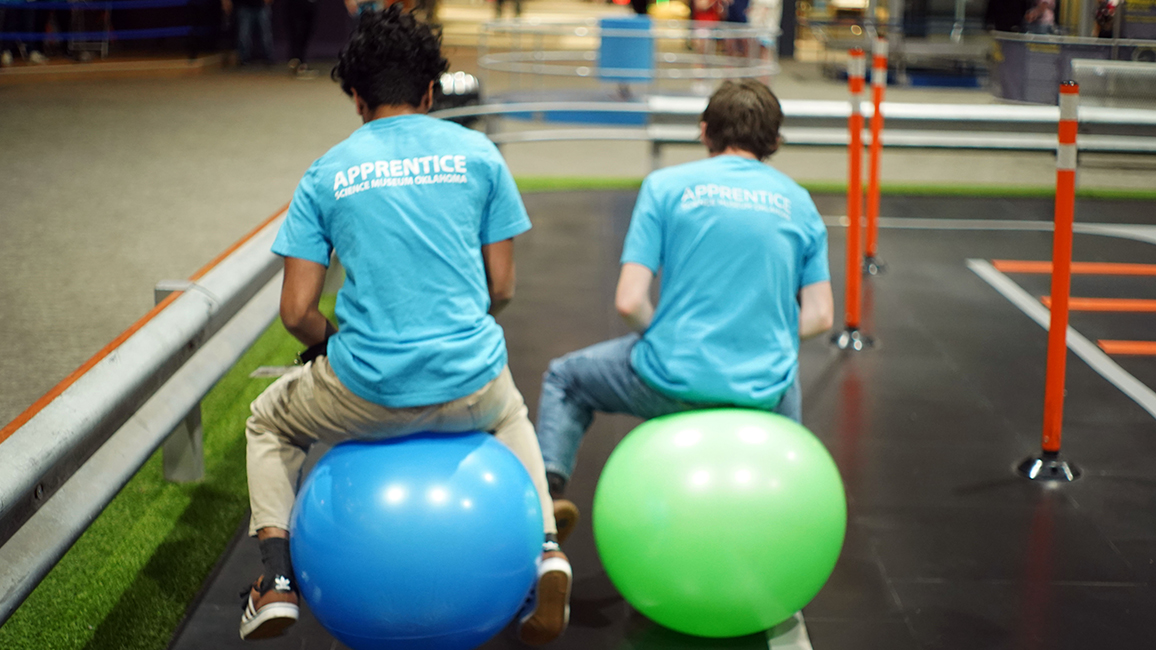 Two teen apprentices in matching blue shirts labeled “Apprentice – Science Museum Oklahoma” bounce on large inflatable balls inside a museum activity zone.