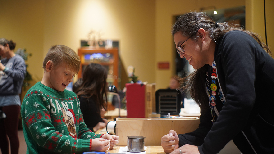 A staff member at Science Museum Oklahoma helps a boy in a chirstmas sweater in Tinkering Garage.