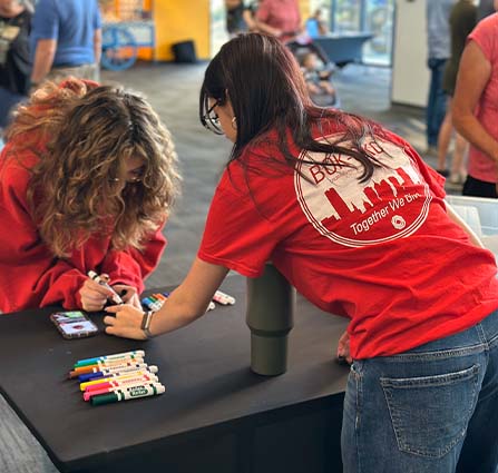 Two volunteers in red shirts decorating items with colorful markers at a table inside Science Museum Oklahoma, with other guests visible in the background.